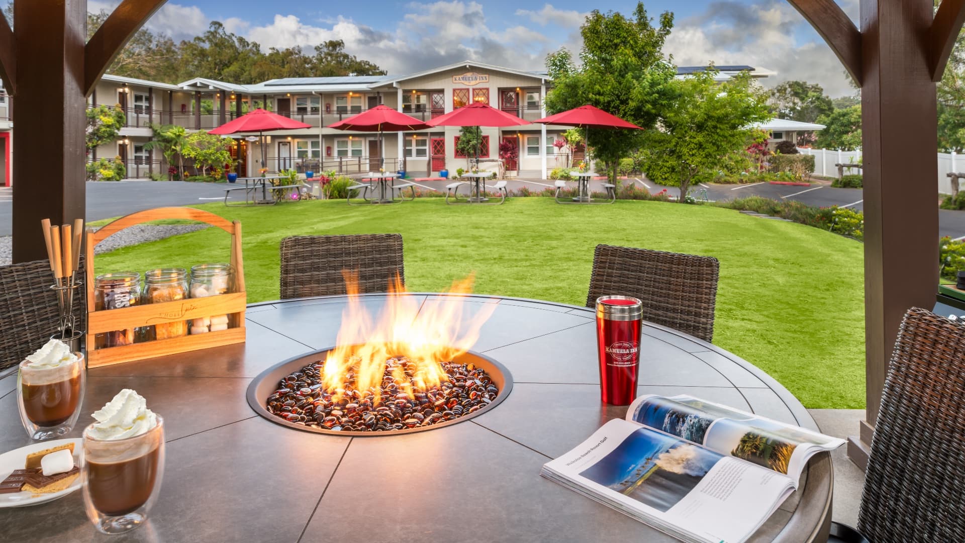 An outdoor seating area with a fire pit, coffee drinks, and a view of a quaint inn surrounded by lush greenery and umbrellas.