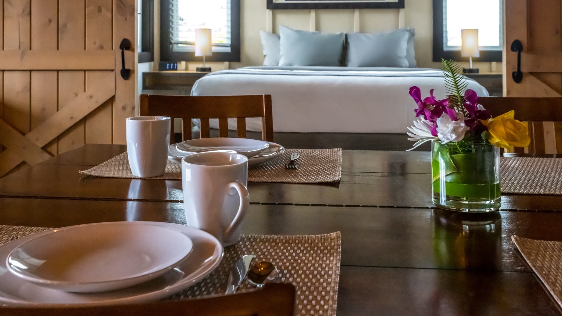 A cozy dining setup with two mugs and plates, facing a well-lit bedroom with a large bed in the background.